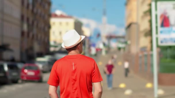 白い帽子の男の観光客は道に沿って通りに沿って晴れた日に歩く — ストック動画