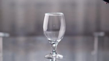 Masadaki boş bardakta kırmızı şarap şişeden dökülüyor, arka planda ışık var.