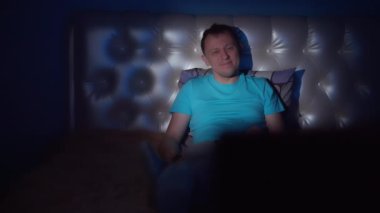 Mavi tişörtlü genç bir adam gece geç saatlere kadar bir yatakta oturur ve televizyon seyreder.