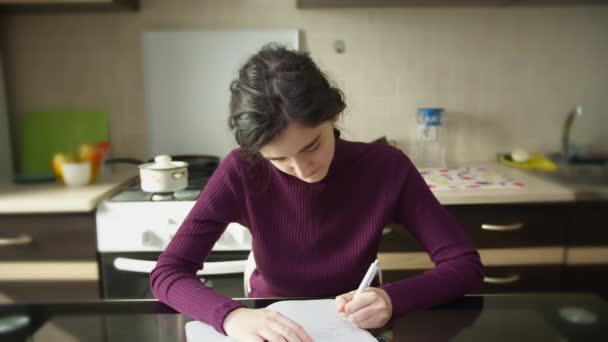 Nettes Studentenmädchen macht sich Notizen in einem Notizbuch und sitzt am Küchentisch — Stockvideo