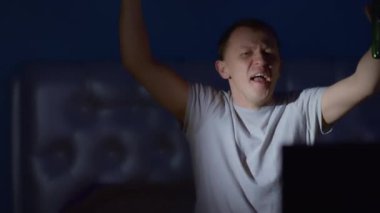 Elinde bira şişesi olan neşeli adam gece TV karşısında oturur, elini kaldırır, kamera hareket eder.