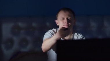 Genç adam gece geç saatlerde televizyon izliyor, kanal değiştiriyor, kamera hareketi yapıyor.