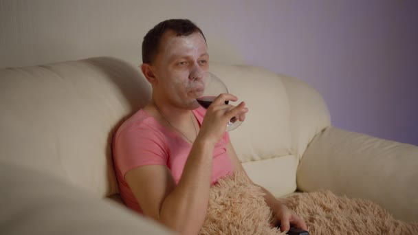 戴着化妆品面具的男人坐在沙发上看电视 喝红酒 — 图库视频影像