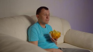 Elinde cips olan genç bir adam akşamları kanepede oturur ve bakar.