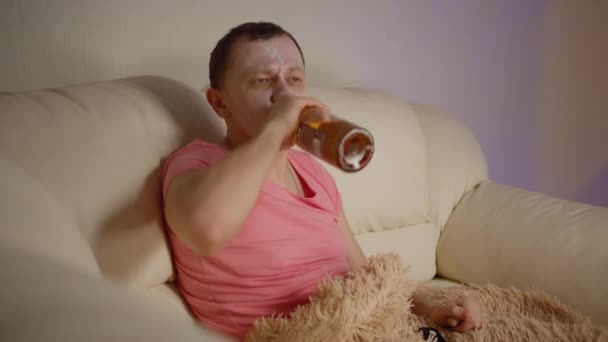 化粧品マスクの若い男がソファの上に座ってテレビを見て アルコールと飲み物のボトルを開きます — ストック動画