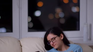 Gece geç saatlerde elinde telefonla bir kanepede oturan gözlüklü bir kız pencerenin arkasına yaslanır.