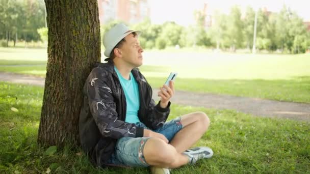 Homme avec chapeau par une journée ensoleillée assis avec un téléphone portable sous un arbre dans un parc de la ville, mouvement de la caméra — Video