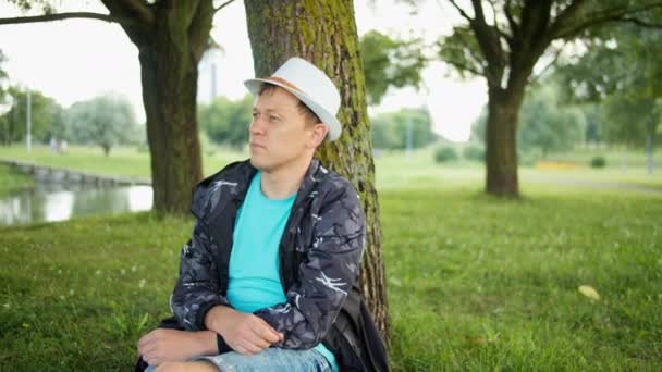 Homem atraente em chapéu descansando sob uma árvore em um parque da cidade, câmera de rastreamento — Vídeo de Stock