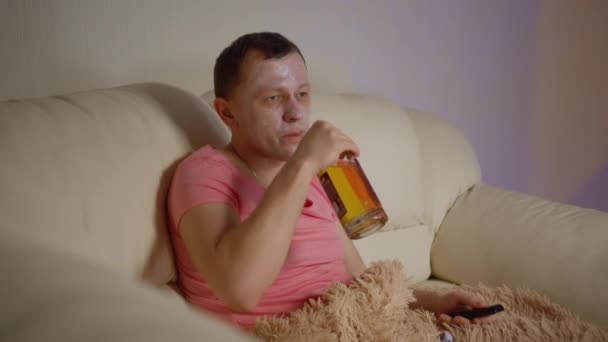 Un hombre con una máscara cosmética en la cara y una botella de alcohol en la mano mira la televisión por la noche — Vídeo de stock