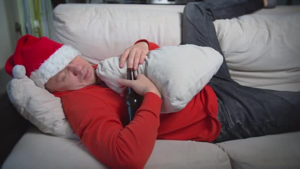 Jeune homme non rasé dans un bouchon rouge dormant sur le canapé tenant une bouteille de bière à son — Video