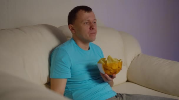 Solitario joven se sienta en el sofá por la noche viendo la televisión y comiendo patatas fritas — Vídeos de Stock