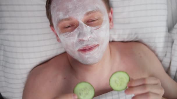 En man ligger med en kosmetisk kräm i ansiktet, framför ögonen gurka skivor, ovanifrån, kamerarörelse — Stockvideo