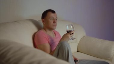 Elinde bir kadeh kırmızı şarapla gülümseyen genç adam geceleri televizyon seyrediyor.