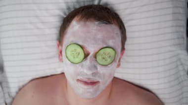 Yüzünde kozmetik maske ve gözlerinde salatalık dilimleri olan gülümseyen genç bir adam.