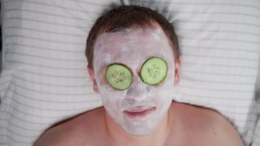 Yüzünde kozmetik maske ve gözlerinde salatalık dilimleri olan gülümseyen bir adam.