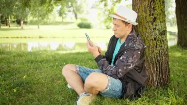 Elinde telefon olan beyaz şapkalı bir adam parkta bir ağacın altında oturuyor. Kamera hareketi, eğlence konsepti.