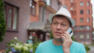 Beyaz şapkalı genç adam sokakta cep telefonuyla konuşuyor, kamera takip ediyor.