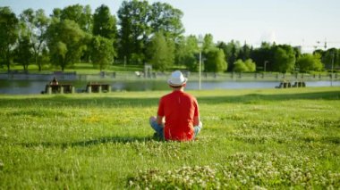 Beyaz şapkalı genç adam çimlerin üzerinde oturur, gölete bakar, kamera hareketi, arka plan