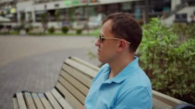 Güneş gözlüklü genç adam kentin bulanık arka planında bir bankta otururken dinleniyor, kamera hareketi
