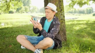 Elinde telefon olan beyaz şapkalı bir adam parkta bir ağacın altında oturuyor, kamera hareketi, eğlence konsepti.