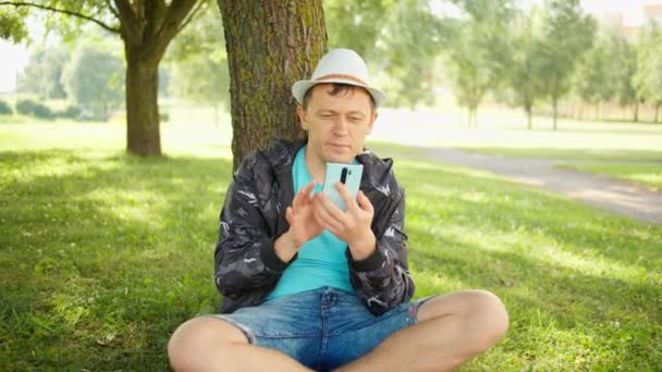 Молодий Чоловік Сидить Під Деревом Парку Телефоном Руках Концепція Відпочинку — стокове відео