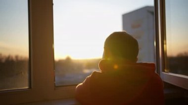 Kapüşonlu bir adam pencerede dikilirken gün batımına bakıyor.