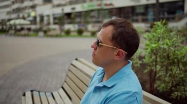 Güneş gözlüklü adam şehrin bulanık arka planında bir bankta otururken dinleniyor.