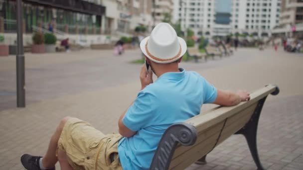 Turista masculino en un sombrero blanco descansa mientras está sentado en un banco, movimiento de la cámara — Vídeos de Stock