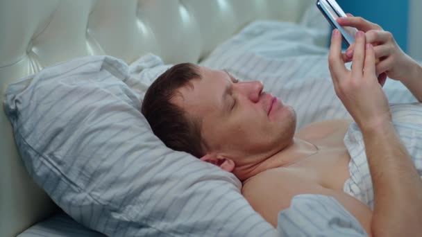 Hombre hojeando noticias en el nuevo teléfono, acostado en la cama, movimiento de la cámara — Vídeo de stock