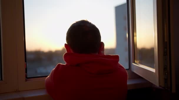 Homem de camisola olha para o pôr-do-sol em pé em uma janela aberta, movimento da câmera — Vídeo de Stock