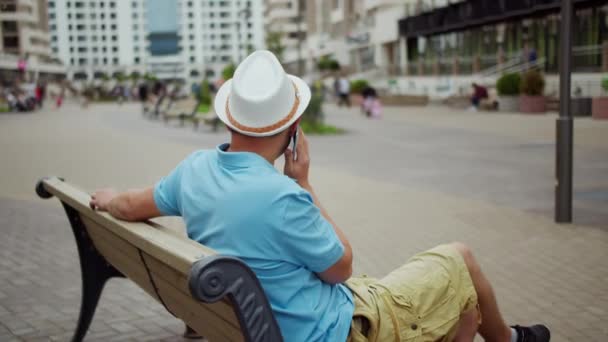 Молодой человек в шляпе говорит по телефону, сидя на скамейке, движение камеры, вид сзади — стоковое видео