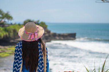 Endonezya, Bali 'de turistlerle dolu bir plajın yanında mavi bir elbise ve şapka giyerek sıcak bir yaz gününün tadını çıkaran tek başına bir bayan yolcu.