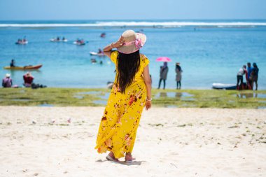 Endonezya, Bali 'de turistlerle dolu bir plajın yanında sarı bir elbise ve şapka giyerek sıcak bir yaz gününün tadını çıkaran tek başına bir bayan yolcu.
