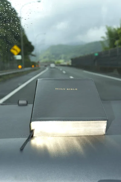 Holy Bible with golden glow. Raindrops on the windshield. Blurred background expressway with cars.