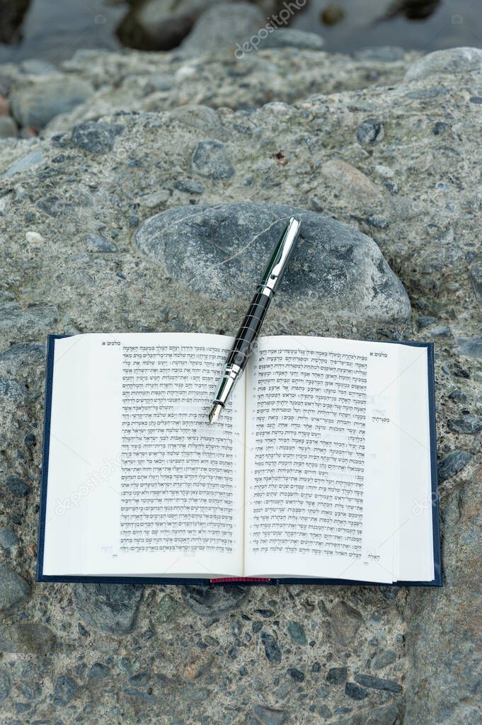 Open Hebrew Bible - TANAKH (Torah, Neviim, Ketuvim - The Law, The Prophets, The Writings) with fountain pen outdoors. Isolated on stones background. Vertical shot. Close-up.