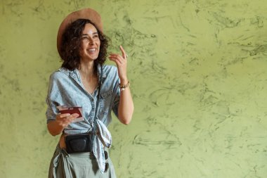 Belgeleri ve uçak biletleri olan kıvırcık saçlı mutlu kız. Kot ceketli ve şapkası duvara dayanmış bir kız.