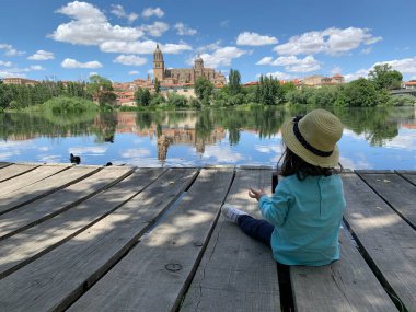 Şapkalı küçük kız nehir kıyısında oturmuş ördekleri besliyor.. 