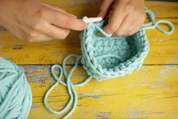 Strick- und Handhäkelkonzept. Frauenhände stricken einen türkisfarbenen Korb mit einem großen Haken an der Kordel. hölzernen gelben Hintergrund. Ansicht von oben mit Kopierraum. — Stockfoto