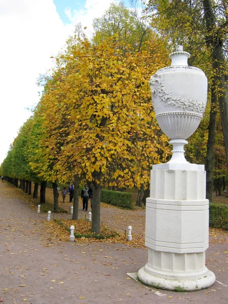 Golden autumn concept. Marble antique vase on a pedestal on the background of linden alley in the autumn park. — Stock Photo, Image