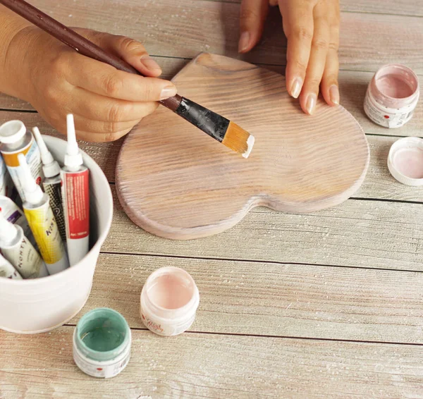 Γυναικεία σχέδια χέρι με ένα πινέλο και ακρυλικά χρώματα σε ένα φύλλο από ξύλο πεύκου θέρμανση.Επίπεδη θέσει με γυναίκες όμορφα χέρια και μοντέρνα χρώματα σε ρουστίκ ξύλινο τραπέζι. αντίγραφο χώρου.Handcraft έννοια ξύλου — Φωτογραφία Αρχείου