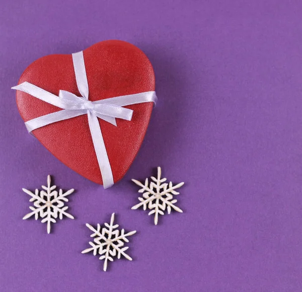 Feliz Navidad y Feliz Año Nuevo tarjeta de felicitación. caja de regalo de color rojo de Navidad en forma de corazón con cinta blanca y copos de nieve sobre fondo púrpura.. —  Fotos de Stock