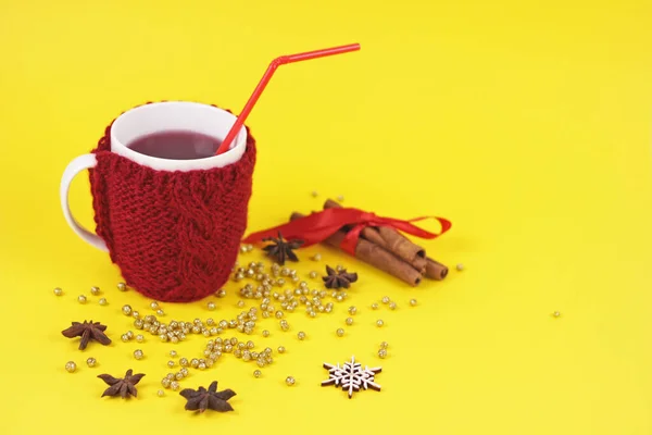 Mulled wine in mug with woolen case on yellow background.cup in beuatiful knitting red case with spices and golden glitters.Christmas New Year greeting card.Warm and cozy hygge holiday concept.