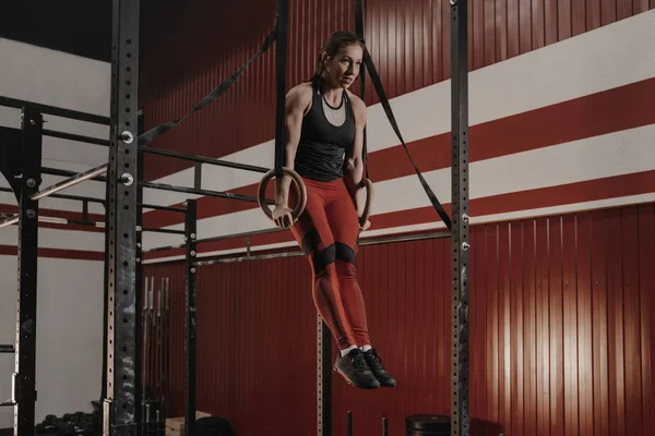 Full body strong sportswoman hanging on gymnastic rings and swinging while  exercising during intense training in dark gym stock photo