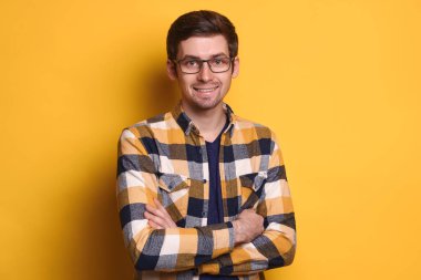 Genç beyaz adamın portresi mutlu ve gülümseyen, gözlük takan ve sarı arka planda izole edilmiş. Kollarını kavuşturup kameraya poz veren yakışıklı öğrenci. İnsan duyguları