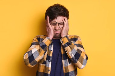 Gözlüklü, baş ağrısından muzdarip sinirli, stresli, yorgun, bitkin ve fazla çalışan genç adamın stüdyo portresi. Sarı arka planda izole edilmiş. Aşırı çalışma, profesyonel yorgunluk, migren.