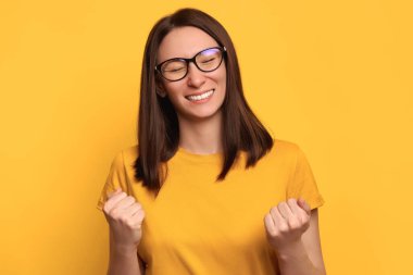 Gözlüklü ve sarı tişörtlü genç esmer kadın sarı arka planda yalnız poz veriyor. Kazanan hareketi yapıyor. İnsanlar samimi duygular ve kazanan konsept. Kopyalama alanı oluştur