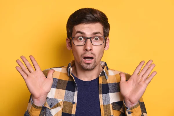 Young amazed handsome man in glasses standing on yellow wall raising arms wondering, astonished to see something, facial expression, shocked with news, amazed. People, reaction and emotions concept