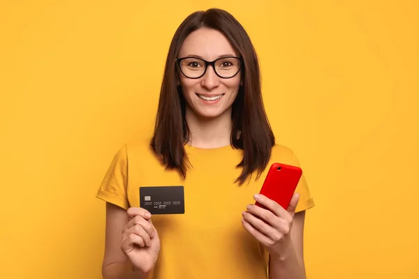Happy beautiful woman in eyeglasses and yellow t-shirt using smartphone and credit card for online shopping in online store, online payment, mobile banking, paying domestic bills. Technology concept