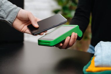 NFC terminali ve giysi dükkanındaki cep telefonu üzerinden ödeme yapan müşterilerin yakın çekimleri. Akıllı telefonla öde. Bağlantısız NFC ve modern cihaz teknolojisi kavramı