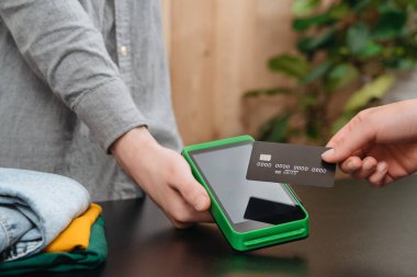 Müşterinin elinde NFC ödeme terminalinin yanındaki banka kredi kartı var. Elinde de giyim mağazasındaki kasiyer. Müşteri kredi kartıyla alışveriş yapar. Bağlantısız, cep telefonu, hızlı ödeme, nFC konsepti 